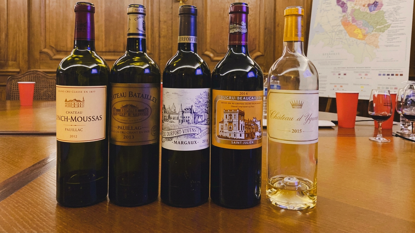 Five bottles of wine displayed on a wooden table, including red and white varieties from various French chateaus. The backdrop features a map of wine regions and red plastic cups are visible.