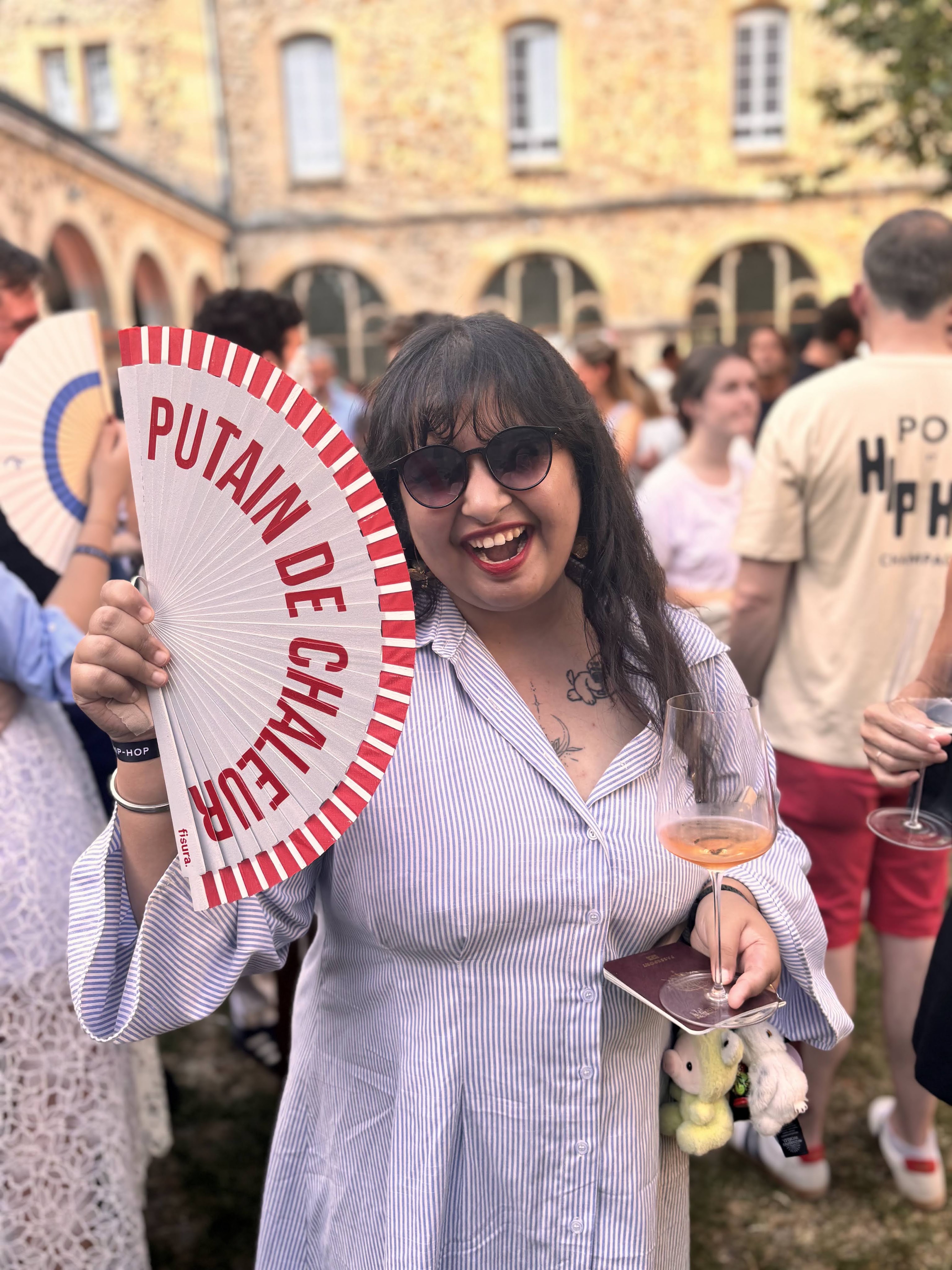 A woman in a striped shirt holds a decorative fan with the words 'PUTAIN DE CHALEUR' while smiling at a social gathering. She also has a glass of rosé wine and small stuffed animals attached to her bag.