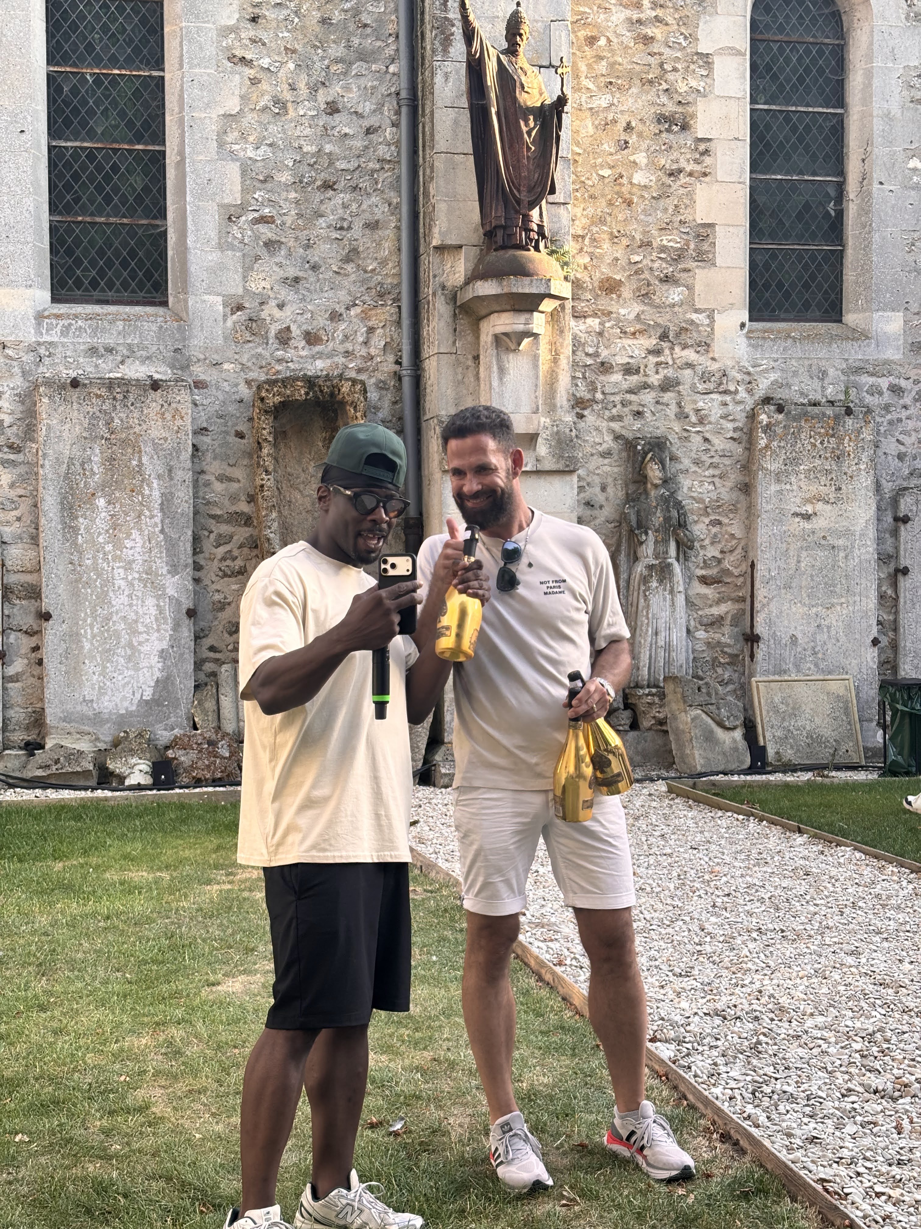Two men standing in front of a stone building, one holding a microphone and a phone, the other holding bottles of drink and giving a thumbs up.