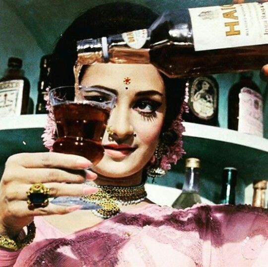 A woman in traditional attire pouring a drink from a bottle into a glass, set against a backdrop of shelves filled with various bottles.