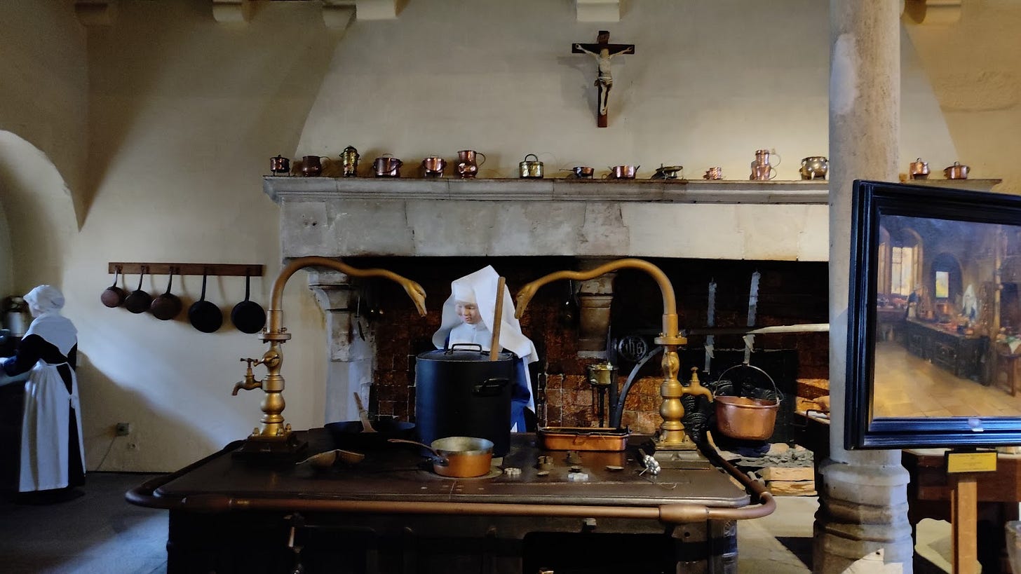 An old-fashioned kitchen with a large stove, pots hanging on a rack, and a fireplace in the background. A person dressed in historical attire is seen near the stove, with kitchen utensils and a framed painting on display.
