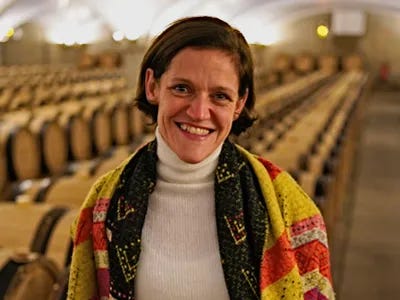 A smiling woman wearing a colorful shawl stands in a wine cellar filled with wooden barrels.