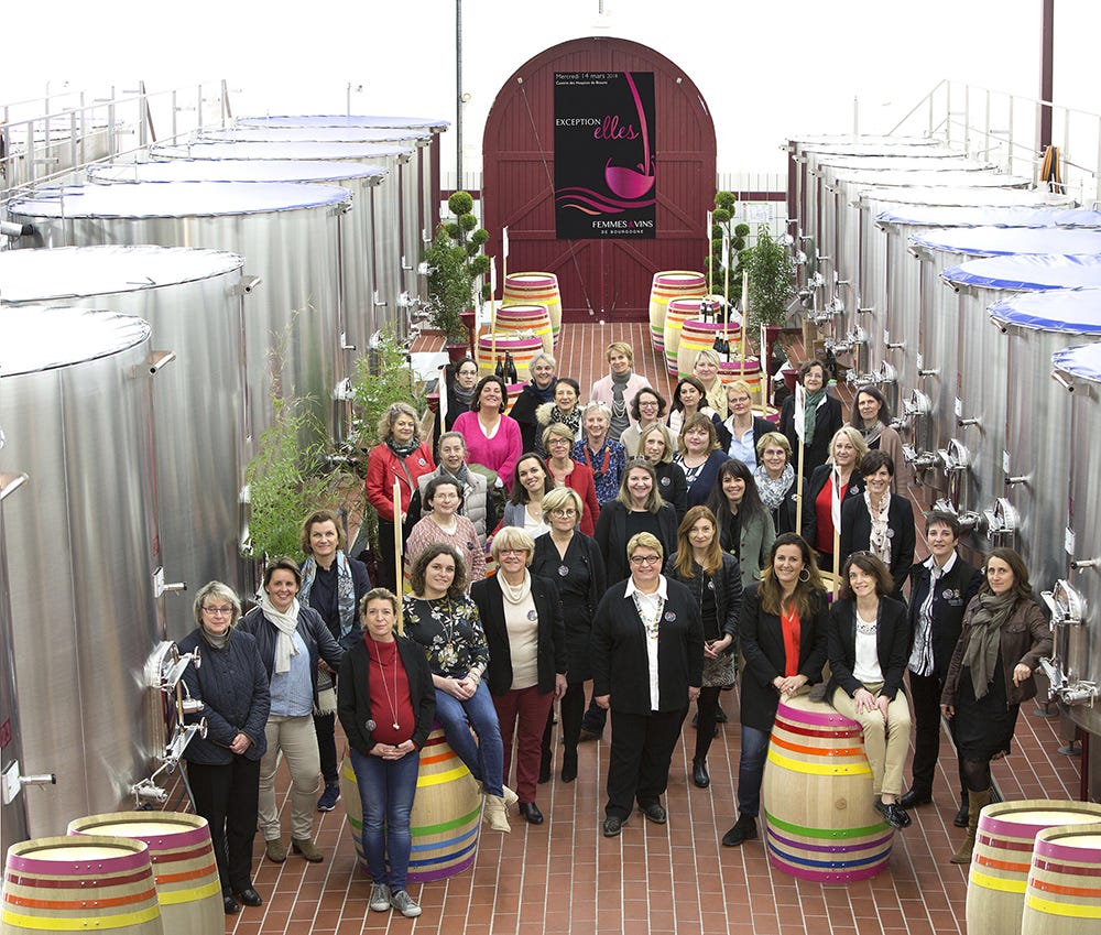 A group of women posing together in a winery, surrounded by metallic fermentation tanks and colorful wine barrels, showcasing a strong community presence in the wine industry.