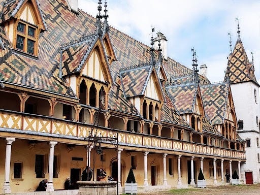 A historical building with a colorful patterned roof, featuring ornate architectural details, wooden beams, and a well in the foreground.