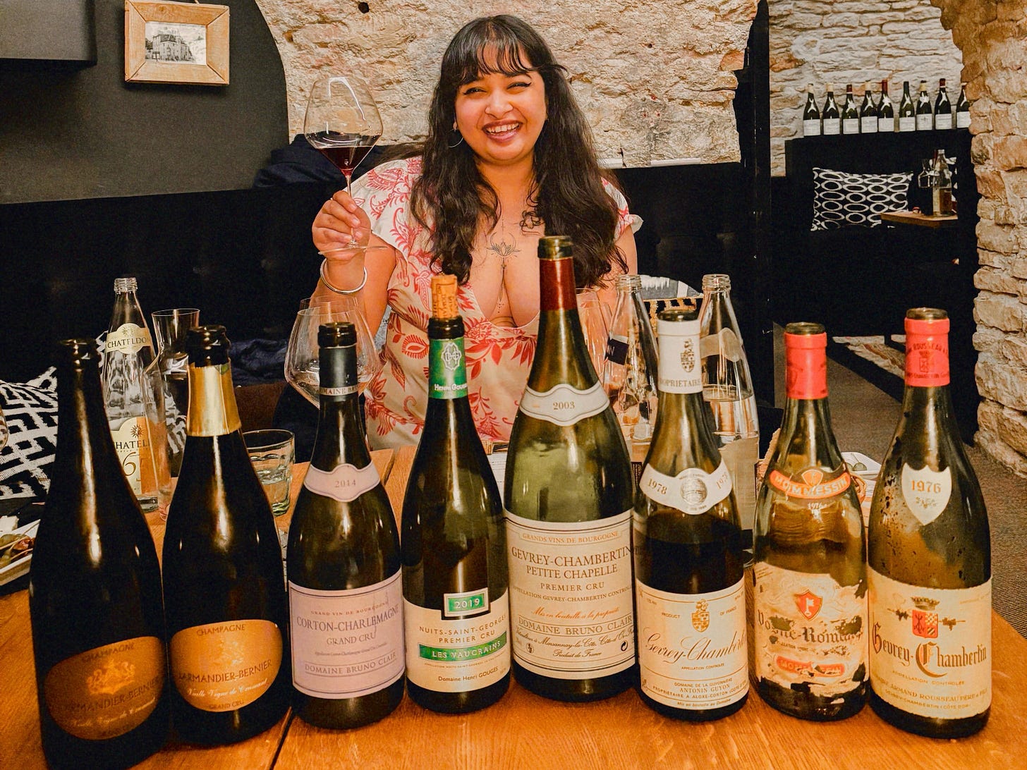 A smiling woman holds a wine glass and raises it, surrounded by a variety of wine bottles on a wooden table.