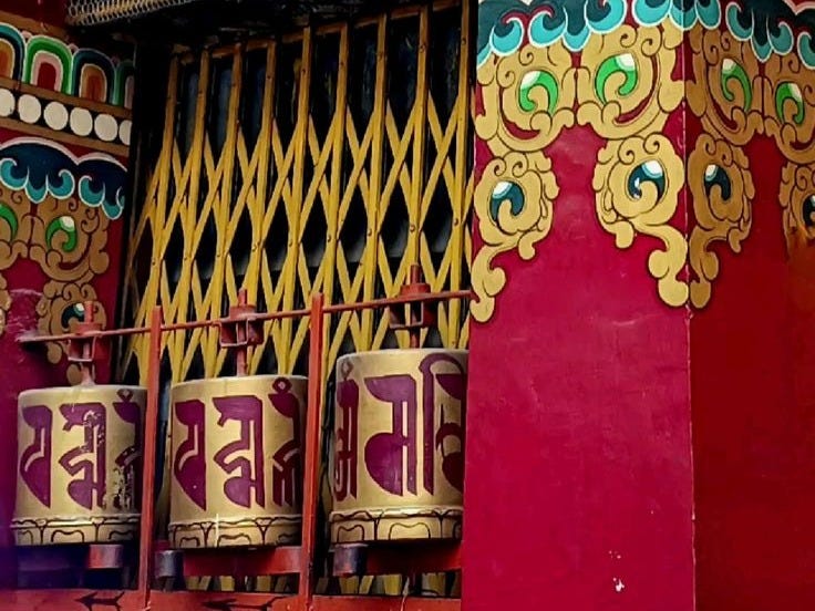 Colorful prayer wheels with inscriptions located near a vibrant temple wall featuring ornate designs and patterns.