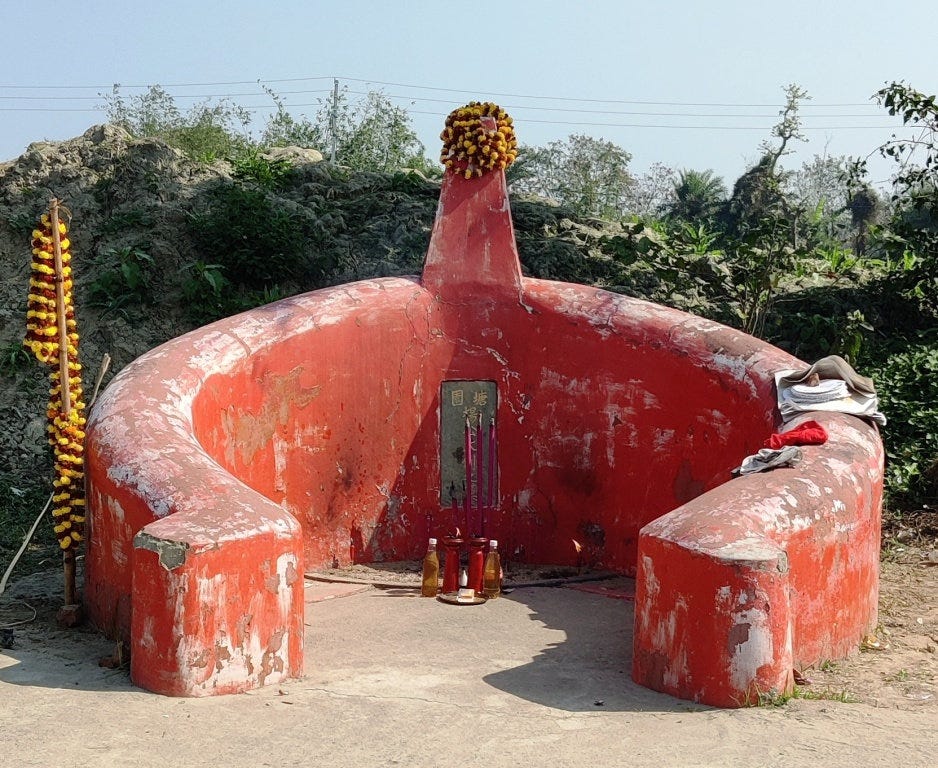 A red, half-circular structure resembling a seat or altar, adorned with marigold flower garlands and incense holders placed in front. The background features greenery and distant trees.