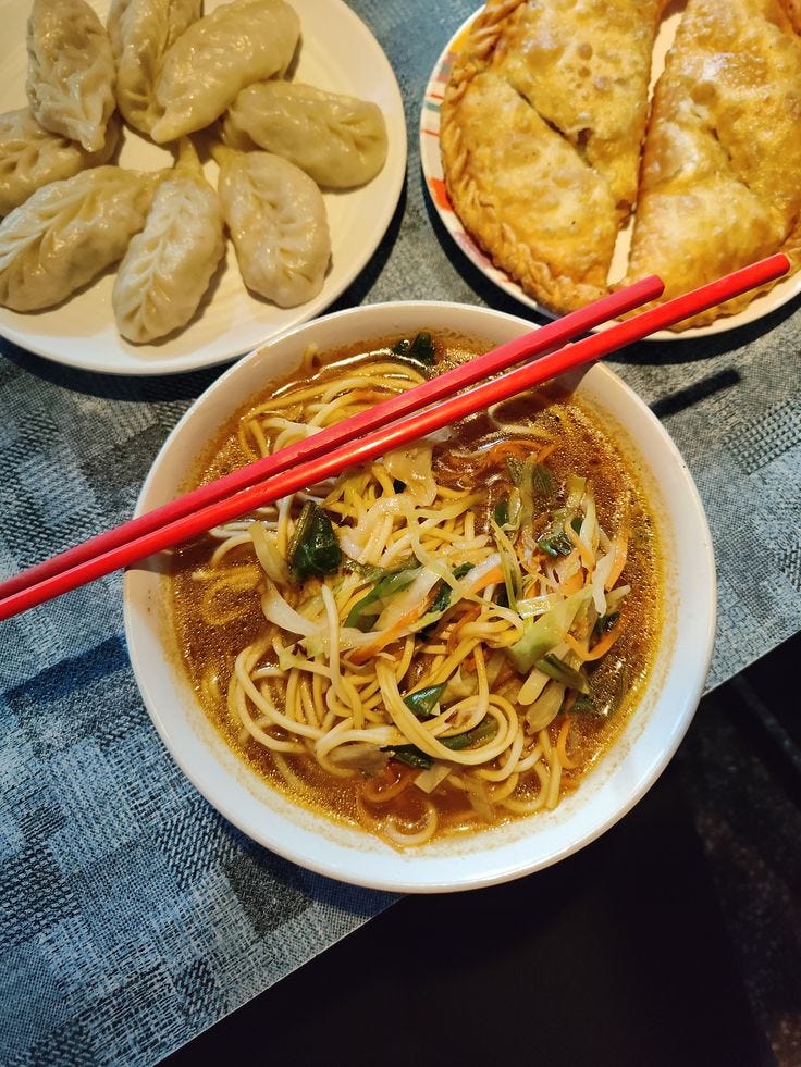 A bowl of noodles topped with green vegetables and served with red chopsticks, alongside plates of dumplings and pastry pockets.