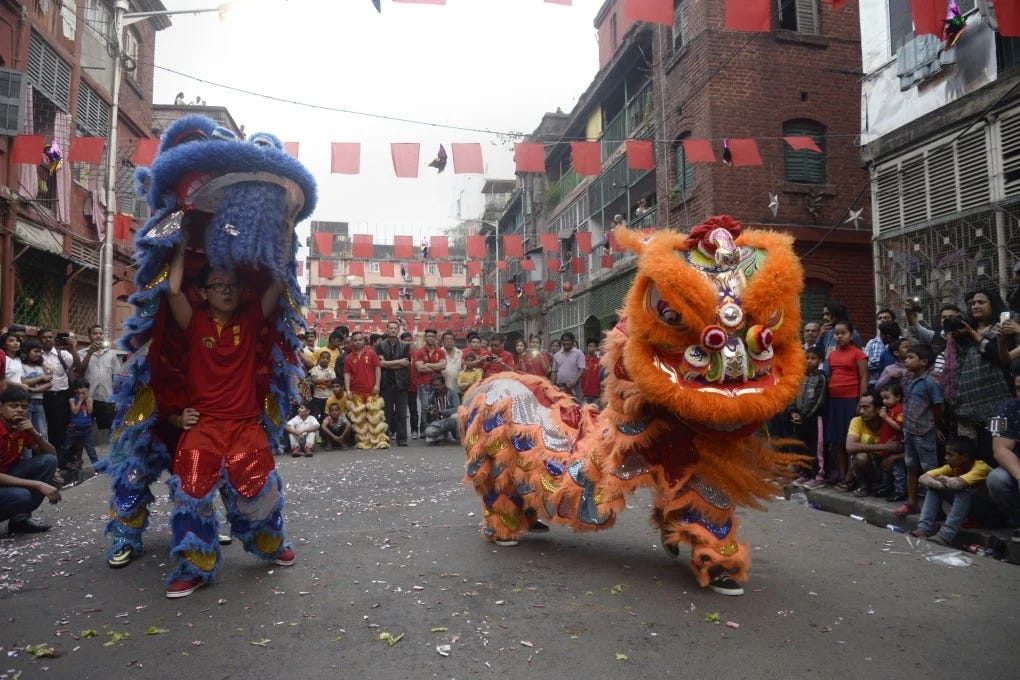 Two performers in colorful lion costumes dance in a street during a festival, with a crowd watching and festive decorations above.