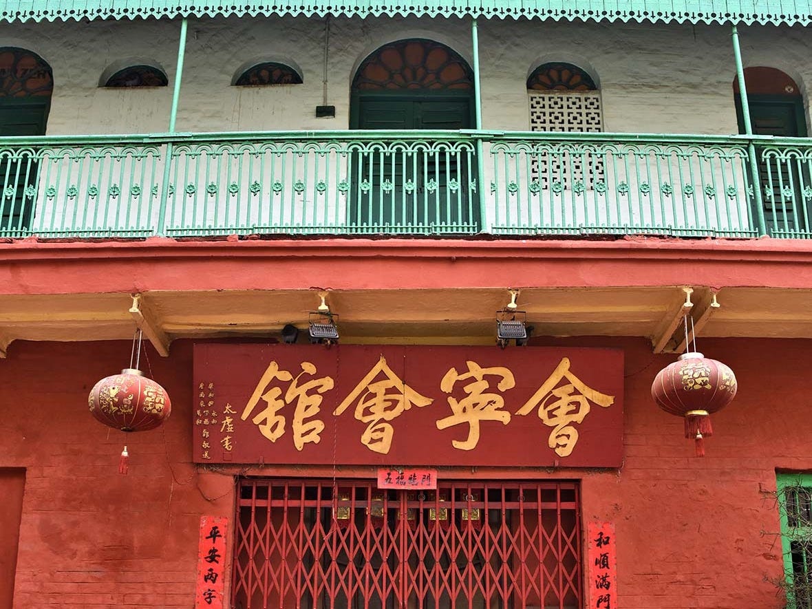 A traditional building facade with red walls, featuring a large sign in Chinese characters. Green balcony with ornate railings is visible above, along with decorative red lanterns hanging from the eaves.