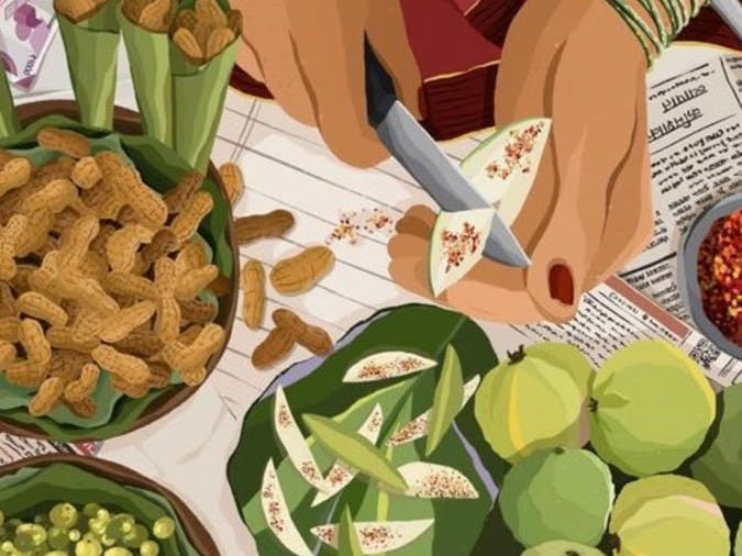 A close-up of a person slicing a green fruit with a knife, surrounded by various ingredients including peanuts, green leaves, and a bowl of red spice, all arranged on a table with newspaper.